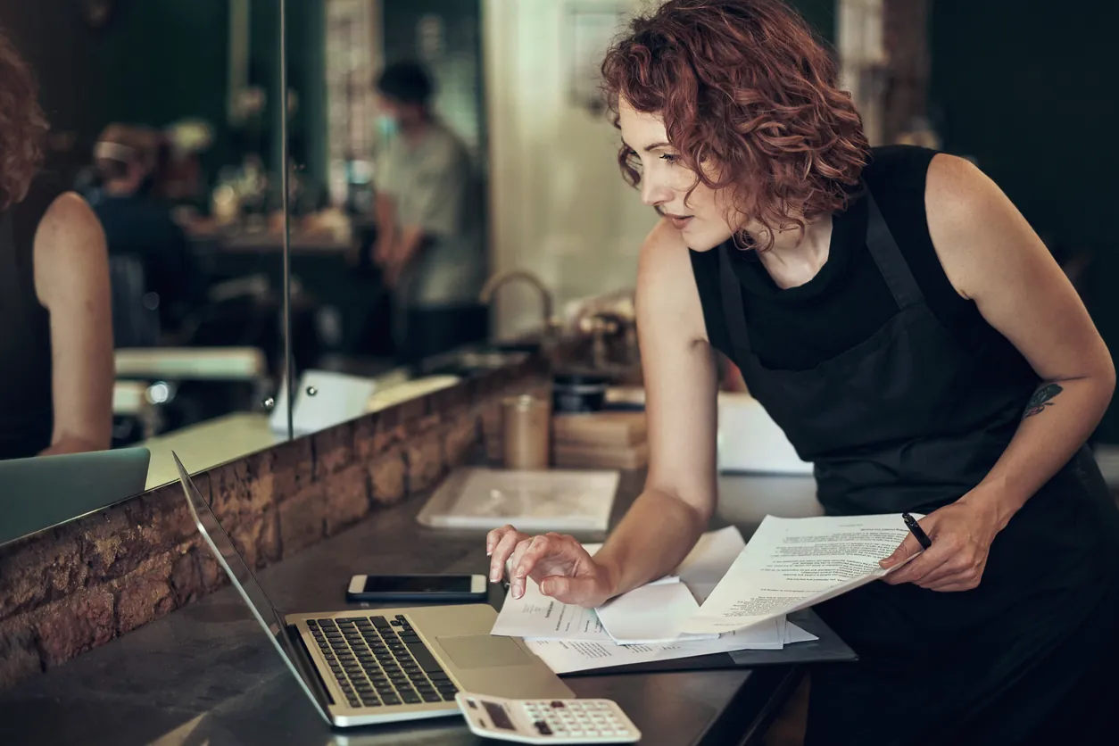 A salon owner at her counter, checking paperwork against her laptop
